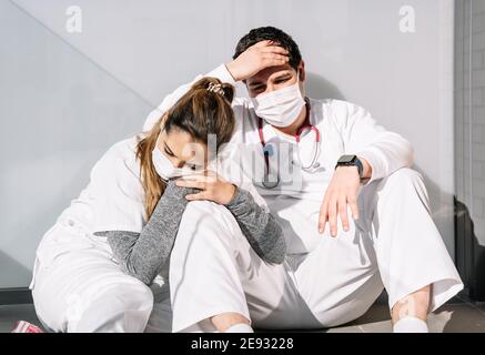 Exhaustos médicos masculinos y femeninos en máscaras protectoras que se apoyan pared de terraza y dormir después de un duro trabajo durante el coronavirus epidemia Foto de stock