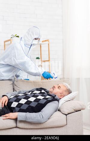 El anciano enfermo la infección, el trabajador médico en traje de avellana examina al paciente y mide la temperatura con termómetro Foto de stock