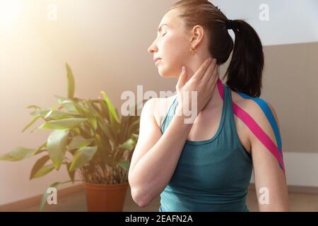Mujeres jóvenes tocando su cuello con la mano y cinta de cinesio elástica en su hombro. Hacer ejercicio en casa. Terapia física de Kinesiología. Foto de stock