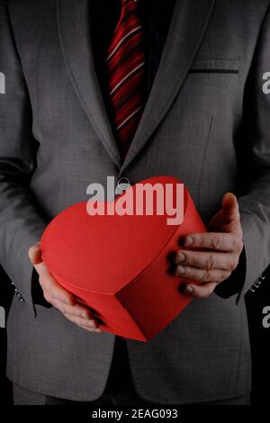 Hombre irreconocible en traje sostiene fuera corazón en forma de caja de regalo rojo, concepto de felicitación, fondo negro Foto de stock