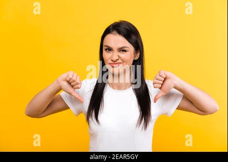 No me gusta el gesto. Una joven caucásica disgustada con camiseta blanca básica muestra un gesto de pulgares hacia abajo y una grimación negativa en su cara, de pie sobre un fondo naranja aislado Foto de stock