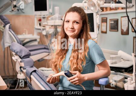Encantadora mujer stomatolgist mirando la cámara mientras sostiene el escalador dental ultrasónico. Dentista femenino sentado en silla en armario de estomatología con equipo especial. Concepto de odontología, salud. Foto de stock