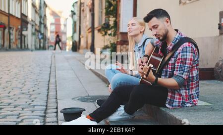 Un par de jóvenes cantantes sentados en la acera, tocando la guitarra y cantando. Concepto de estilo de vida. Foto de stock
