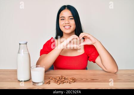Niña asiática bebiendo leche saludable de almendra sonriendo en amor haciendo corazón símbolo forma con las manos. Concepto romántico.