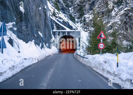 Entrada de un túnel iluminado a lo largo de un camino alpino nevado en invierno. Una señal de carretera que advierte de la caída de rocas en el lado derecho de la carretera.