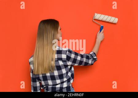 Mujer joven rubia en la camisa de Lumberjack está pintando la pared naranja con rodillo de pintura. Vista posterior. Tiro de cintura arriba.