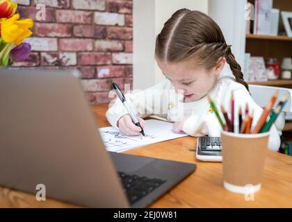 Linda chica joven haciendo cálculos complejos de escritura de matemáticas en escuela en casa con ordenador portátil y calculadora en un escritorio colorido Foto de stock