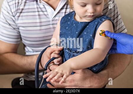 Vacunación de una niña en los brazos de su padre en el consultorio del médico desde el coronavirus. El divertido yeso adhesivo de los niños. Vacuna contra el covid-1