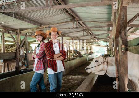 un niño y una niña con ropa informal y un sombrero de pie hacia atrás con las manos cruzadas en un granja de vacas estable