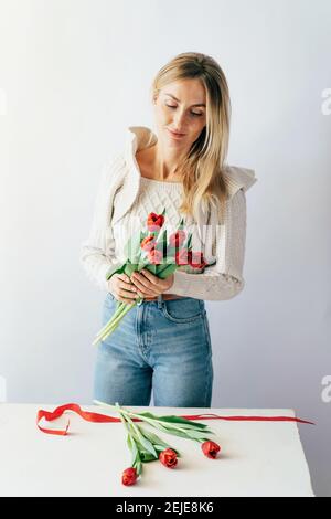 Atractiva mujer joven rubia madura en un suéter y jeans hace un ramo de tulipanes rojos. Foto de stock