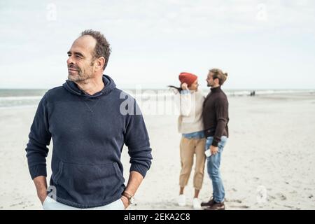 Retrato de hombre de pie en la playa de arena con una pareja joven en el fondo