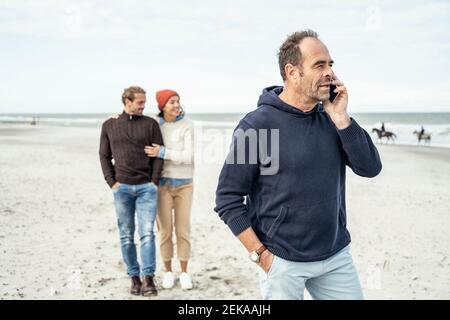 Retrato del hombre de pie en la playa de arena y hablando teléfono inteligente con pareja joven de fondo