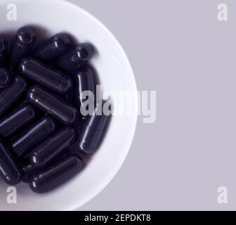 Macro top shot de cápsulas de carbón activado apiladas en un pequeño tazón blanco, sobre un fondo gris violeta. Foto de stock