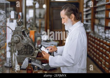 Vista lateral del farmacéutico profesional concentrado de hombre mayor utilizando la tableta digital, trabajando en la farmacia antigua con el interior de la vendimia Foto de stock