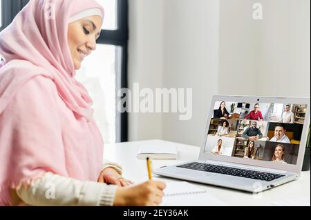 Volver ver mujer musulmana usando hijab usando la aplicación para portátil para la conexión de vídeo con diversos equipos de trabajo. Una estudiante islámica estudiando en línea, viendo un seminario web, tomando notas. Estudio electrónico, videollamada