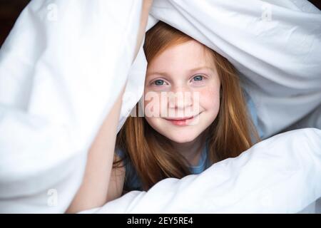 Joven niña de pelo rojo peeking fuera de bajo un edredón blanco. Foto de stock