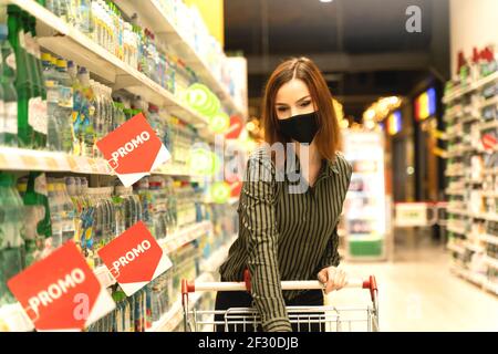 Mujer con máscara en el supermercado hace compras de comestibles. Cliente con carro en hipermercado. Chica de compras en la tienda. Compras seguras