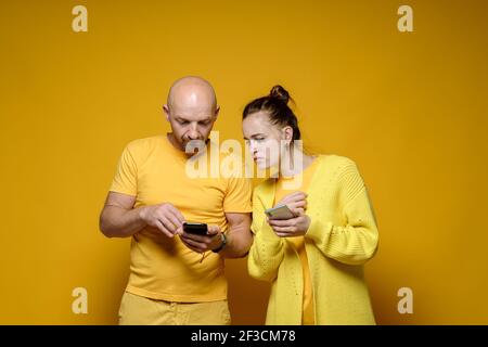 Hombre y mujer serios están sosteniendo smartphones en sus manos y mirando ansiosamente a la pantalla. Fondo amarillo.