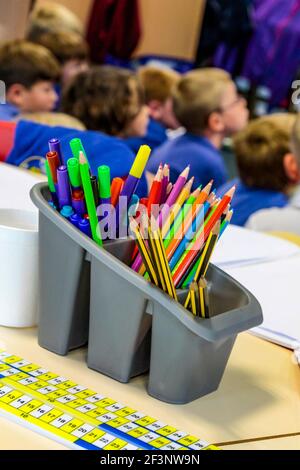 Soporte de plástico gris que contiene plumas y lápices con niños de escuela primaria en un salón de clases al fondo. Foto de stock
