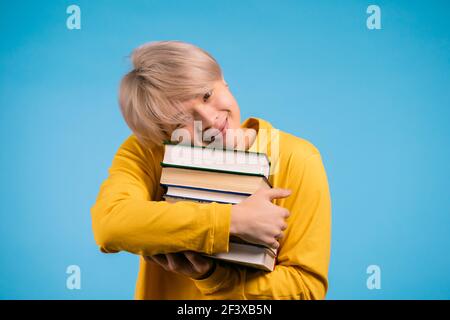 Estudiante coreano de ropa amarilla sostiene una pila de libros universitarios de la biblioteca sobre fondo azul en el estudio. El chico sonríe, está feliz de graduarse. Foto de stock