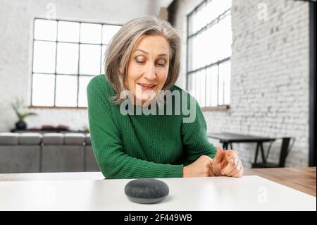 Mujer bonita madura con pelo gris en jumper verde hablando con el asistente virtual digital en casa, haciendo una pregunta o solicitando cambiar de música. Concepto de altavoz Smart AI y control de comandos de voz Foto de stock