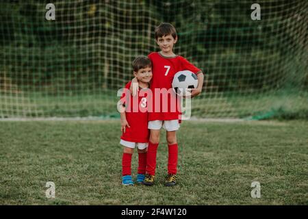 Jóvenes jugadores de fútbol de pie delante de la red de portería. Retrato completo de dos compañeros de equipo de fútbol con vestidos rojos sujetando el balón abrazándose unos a otros.