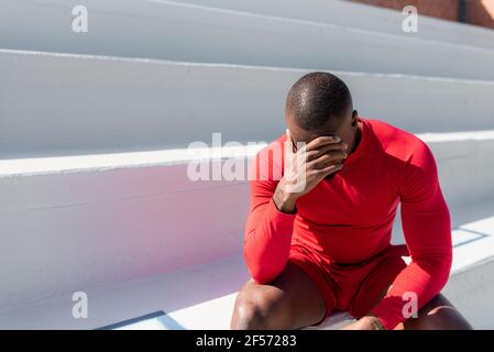 Hombre negro atlético en sportswear sentado en las escaleras cubrir la cara con la mano y mirar hacia abajo.