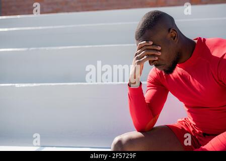 Hombre negro atlético en sportswear sentado en las escaleras cubrir la cara con la mano y mirar hacia abajo.