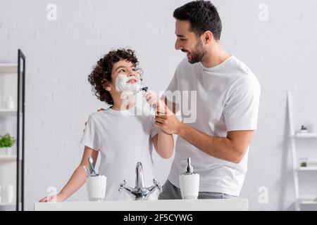 Un hombre musulmán sonriente ayudando a su hijo en espuma de afeitar con una navaja de afeitar Foto de stock