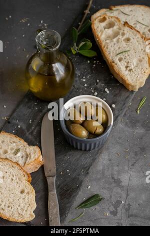 Pan de ciabatta en rodajas italiano en tabla de cortar con hierbas, aceite virgen extra y aceitunas marinadas en fondo grunge oscuro