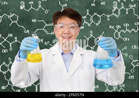 Un científico asiático de medio año que lleva guantes y sostiene un contenedor experimental delante de un pizarrón con una fórmula escrita en él. Foto de stock