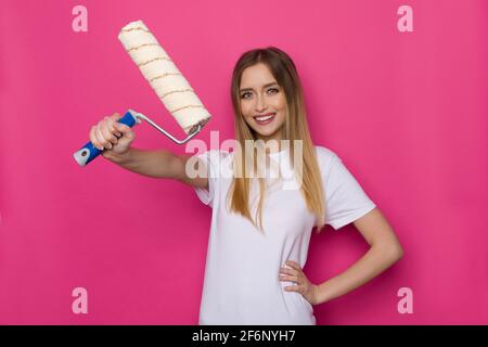 Mujer joven informal segura en camisa blanca está sosteniendo el rodillo de la pintura en una mano y sonriendo. Estudio de cintura arriba sobre fondo rosa. Foto de stock