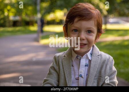 Retrato de un lindo chico de ojos azules, pelirrojo, con un blazer gris y una camisa blanca en un parque en un día soleado