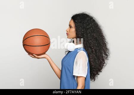 Vista de perfil de una joven negra en ropa de ocio con cabello rizado largo y auriculares alrededor de su cuello sosteniendo un baloncesto en la palma de la mano Foto de stock