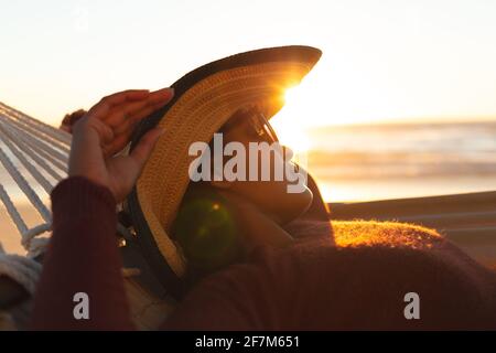 Mujer mixta de carreras en vacaciones en la playa tumbada en una hamaca relajante durante la puesta de sol
