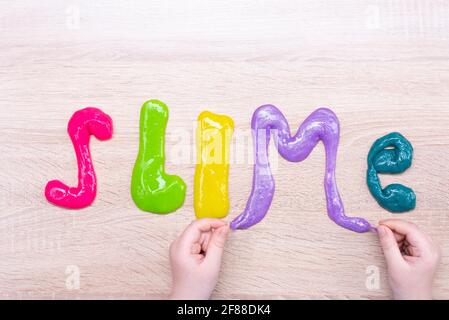 La palabra slime hecho de slimes multi-coloreados en una mesa de madera. Texto en limo de color slime sobre una superficie de madera. Las manos y la limadura del niño. Foto de stock