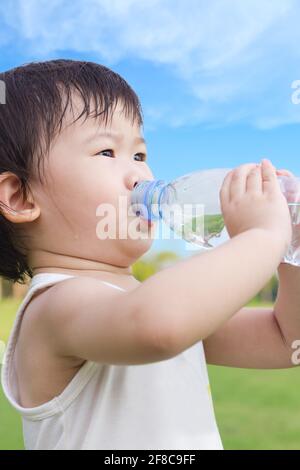 Pequeña niña asiática bebiendo agua de una botella de plástico, después de cansada de un vagabundo en el parque, bajo la luz del sol brillante y el fondo del cielo azul, sho al aire libre Foto de stock