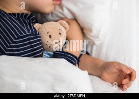 Retrato auténtico enfermo lindo caucásico pequeño niño preescolar en sueño azul con oso de peluche en la cama blanca. Niño descansando a la hora del almuerzo. Cuidado