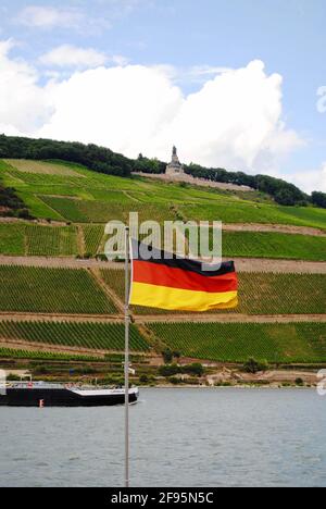 Bingen am Rhein, Alemania: Río Rin con un barco, bandera alemana y viñedos. Niederwalddenkmal, estatua de Germania, un monumento para la reunificación alemana. Foto de stock