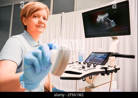 Mujer médica en guantes estériles con transductor de ultrasonidos. Sonógrafo médico femenino que utiliza un moderno ecógrafo en un gabinete ginecológico. Concepto de atención sanitaria y diagnóstico por ultrasonidos. Foto de stock
