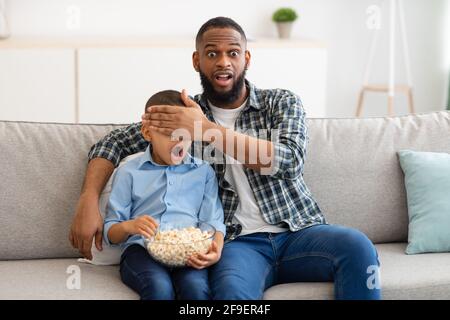 El padre conmocionado que cubre los ojos de su hijo viendo contenidos de televisión cuestionables en interiores Foto de stock