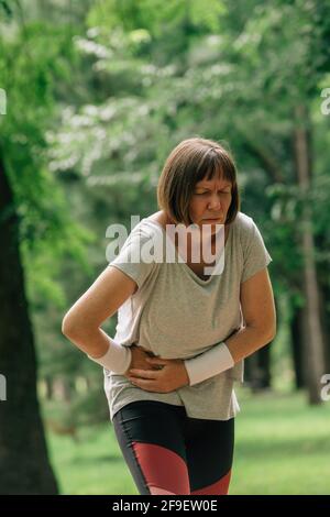 Mujer jogger con dolor de cara grimace después de sentir dolor en abdomen inferior durante el funcionamiento en el parque Foto de stock