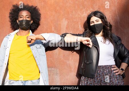 Un par de amigos multiétnicos que llevan máscaras protectoras se saludan unos a otros golpeando los codos y manteniendo la distancia recomendada a evitar Virus Covid-19