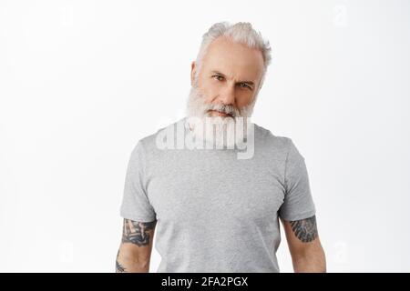 Elegante hombre maduro con cuerpo tatuado y barba larga, inclinación de la cabeza y aspecto de macho seguro en la cámara, mirando a la verdad, de pie en una camiseta gris Foto de stock