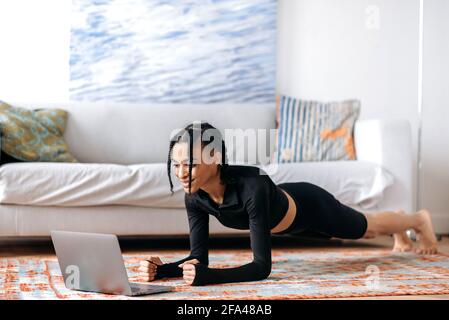 Hermosa joven afroamericana en ropa deportiva, haciendo una tabla en el suelo en casa, usando un ordenador portátil, viendo un vídeo de ejercicios o clases de yoga, llevando un estilo de vida saludable, sonriendo Foto de stock