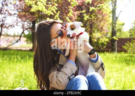 Atractiva mujer joven hipster en gafas de sol besos gato perrito russell terrier en el parque, césped verde y follaje de fondo. Divertido perro de pura sangre besado por Foto de stock