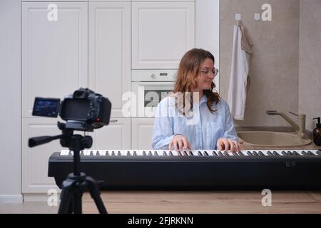 Una mujer graba un vídeo en la cámara mientras ella toca en la cocina de la casa en un piano eléctrico Foto de stock