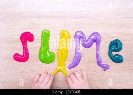 La palabra slime hecho de slimes multi-coloreados en una mesa de madera. Texto en limo de color slime sobre una superficie de madera. Las manos de los niños hacen un insulto Foto de stock