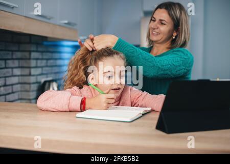 La madre caucásica está arreglando el pelo de la muchacha mientras ella está teniendo clases en línea con una tableta