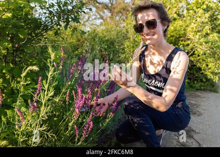 Retrato de joven adulto feliz mujer caucásica recolección hierbas salvia flores silvestres bouquet en el prado al aire libre. Mujer sonriente en monos Foto de stock